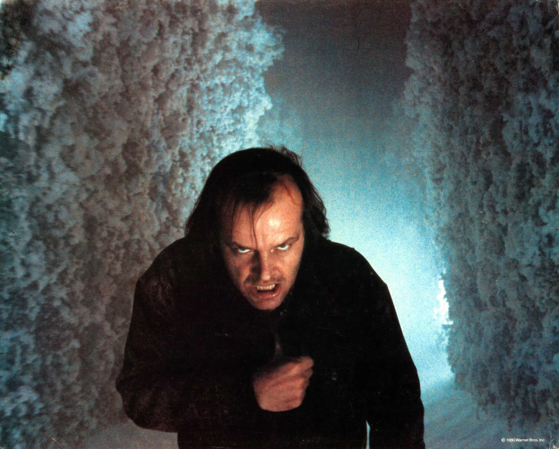 Jack Nicholson como Jack Torrance em 'O Iluminado', com um sorriso maníaco e o rosto enfiado em uma porta quebrada.