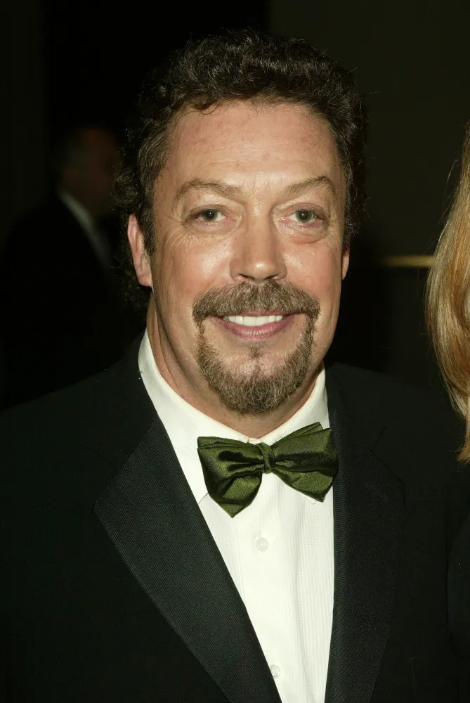 Tim Curry sorrindo de forma travessa, mostrando a personalidade excêntrica que leva para seus personagens.