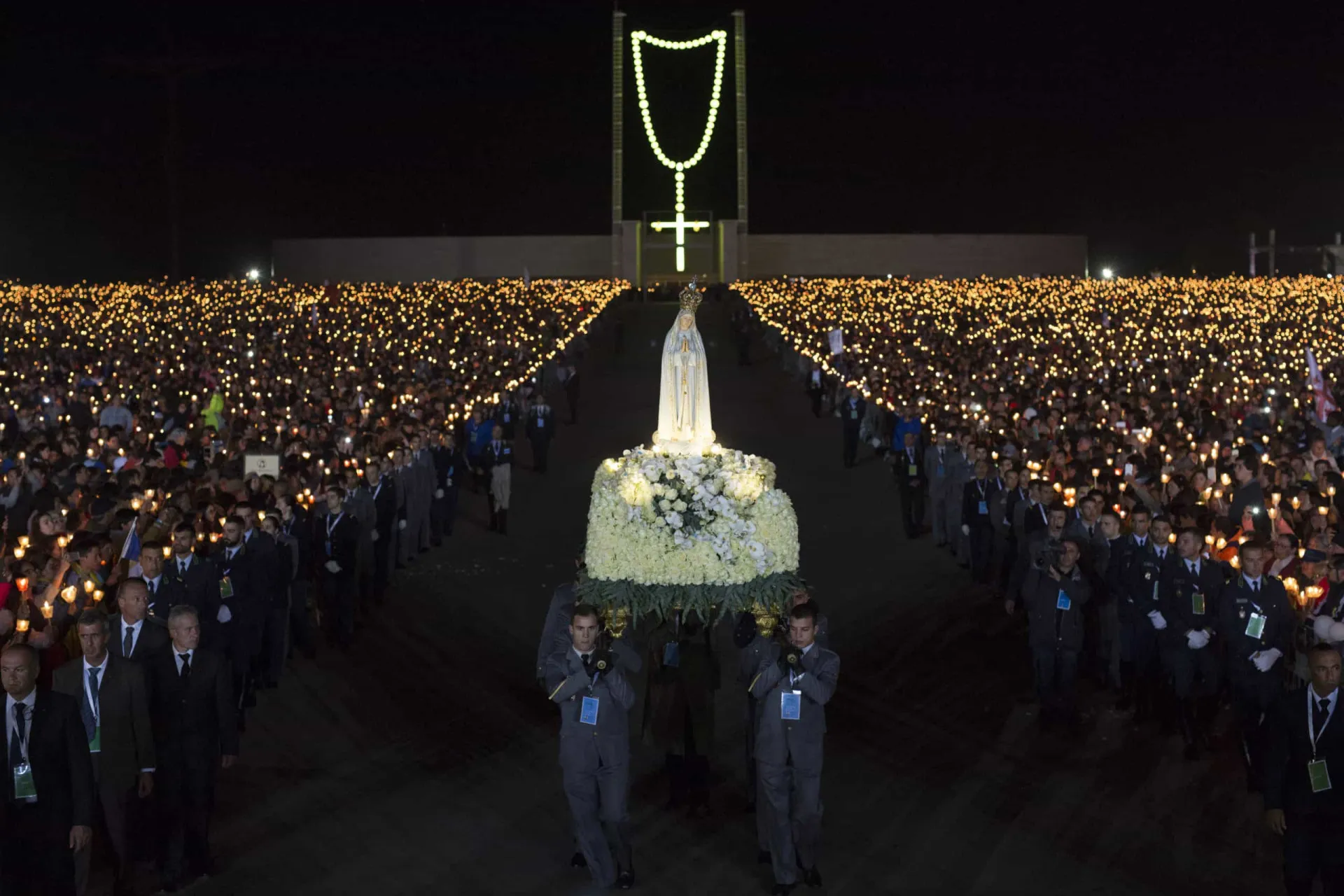 Vista noturna do Santuário de Fátima iluminado, com uma multidão de peregrinos segurando velas.