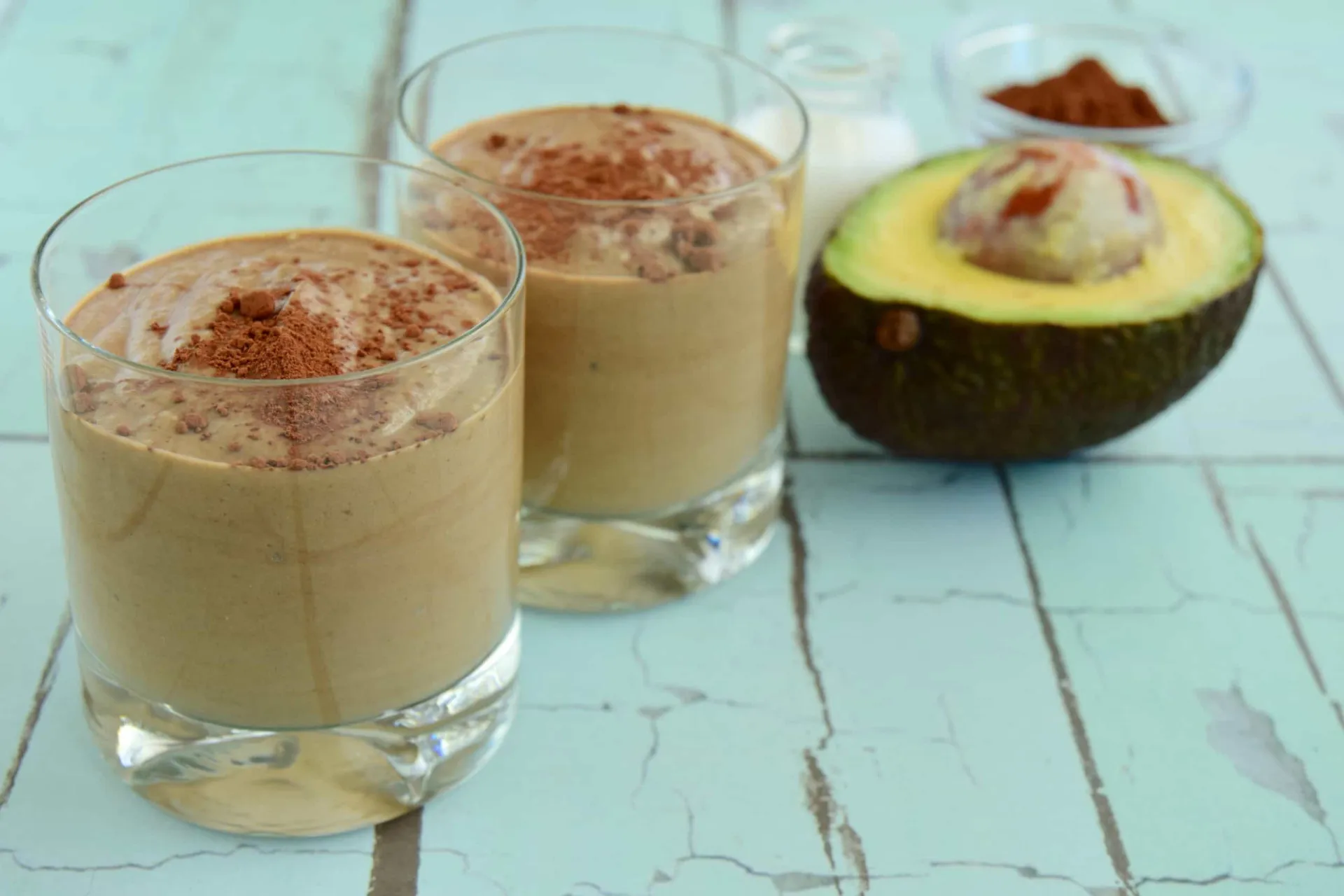 Smoothie espesso e escuro de chocolate e abacate em um copo, com um abacate e chocolate ao fundo.
