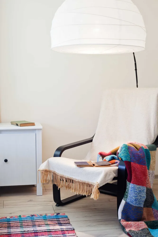 Canto de leitura com uma poltrona confortável, uma luminária de pé, uma manta e uma pilha de livros.